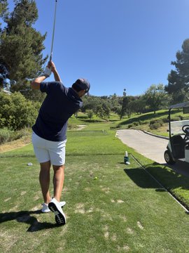The image shows a man swinging a golf club at a ball, likely during a game of golf. He is standing on a green field with his feet firmly planted on the ground as he prepares for his swing. The background features trees and a golf cart parked nearby, indicating that this scene takes place in an outdoor golf course setting.