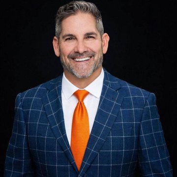A confident, smiling middle-aged man with salt-and-pepper hair and a beard, dressed in a blue windowpane check suit with a white shirt and bright orange tie, poses against a black background—matching Grant Cardone's bold, investor persona as he announces Cardone Capital's expanded Bitcoin allocation for its Fort Lauderdale real-estate project.