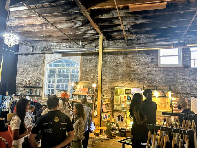 An indoor event space with exposed brick walls, industrial ceiling, and chandelier, where people are gathered among art displays and clothing racks at what appears to be a market or pop-up event.