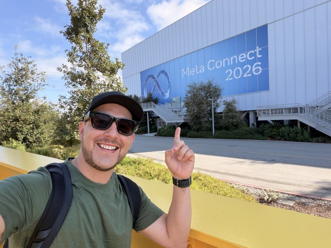 A smiling man in sunglasses, a black cap, and an olive shirt takes a selfie while pointing at a building displaying a 'Meta Connect 2026' sign.