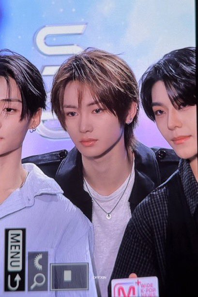 Three young men pose together at a K-pop event, with one wearing a black jacket and silver necklace in the center.