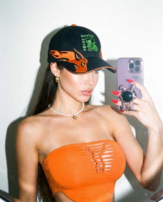 A mirror selfie of a woman wearing an orange cut-out top, a pearl necklace, and a black 'Harry Potter Quidditch' trucker hat with orange flame trim, holding a phone with a pop socket.