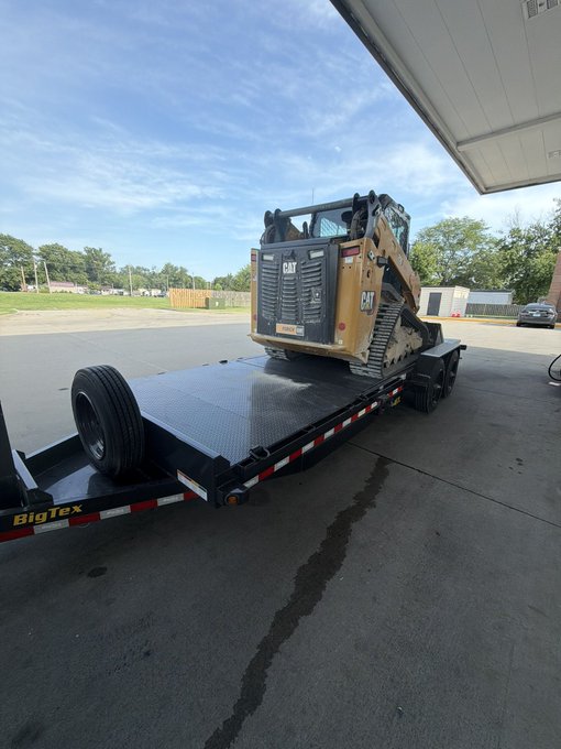 A yellow Caterpillar skid steer loader secured on a black "BigTex" flatbed trailer, parked under a covered area at what appears to be a fuel station.