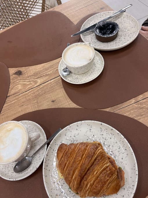 Overhead view of a cafe table with two cappuccinos, a chocolate lava cake, and a large croissant on speckled ceramic dishware.