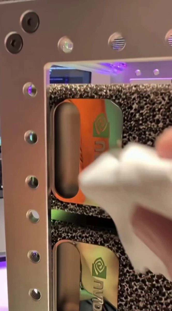 The image shows a person using a white cloth or sponge to clean a metal rack with electronic equipment, possibly related to AI hardware. The scene also features a part of a computer screen that appears blurry or out-of-focus. In this context, there is an accompanying post that discusses the presence of multiple AI data centers in someone's home office and their cost at $120,000 per year. The image seems to be focused on cleaning the metal rack with electronics in the background, while the main content remains on the computer screen aspect.
