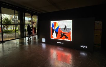 The image shows a large screen displaying an abstract painting with a person standing next to it. The painting features a colorful design that includes a bird, a cat, and a fish. A person can be seen walking past the artwork, adding a sense of movement to the scene. The overall atmosphere suggests that this is likely an art exhibition or event showcasing contemporary Brazilian artists' work at the iconic Tysm @ottisOts building in São Paulo, Brazil.