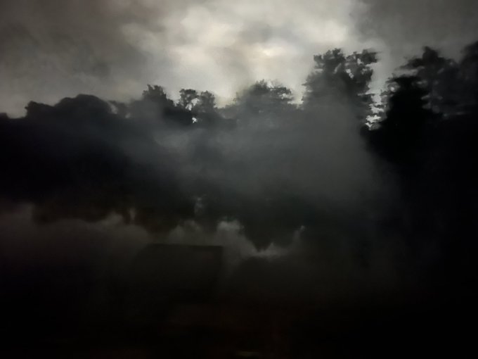 A dark, moody landscape photo showing silhouetted trees reflected in water under a cloudy night sky.