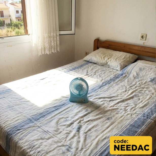 A bedroom with a small blue portable fan on the bed serves as the "air conditioning" from a summer rental listing, humorously highlighting the contrast between advertised amenities and reality, with a yellow promotional overlay displaying "code: NEEDAC" for a Decibel trading giveaway offering a real AC unit.