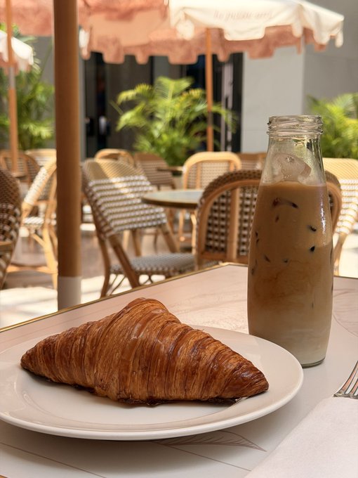 A freshly baked croissant served on a white plate next to a glass bottle of iced coffee at an outdoor cafe with peach-colored umbrellas and woven chairs.