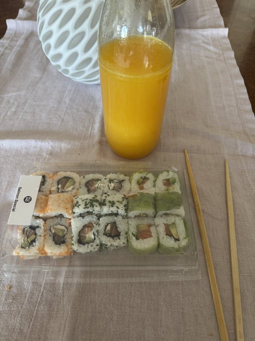A plastic tray containing an assortment of sushi rolls accompanied by bamboo chopsticks and a bottle of orange juice, set on a striped tablecloth.