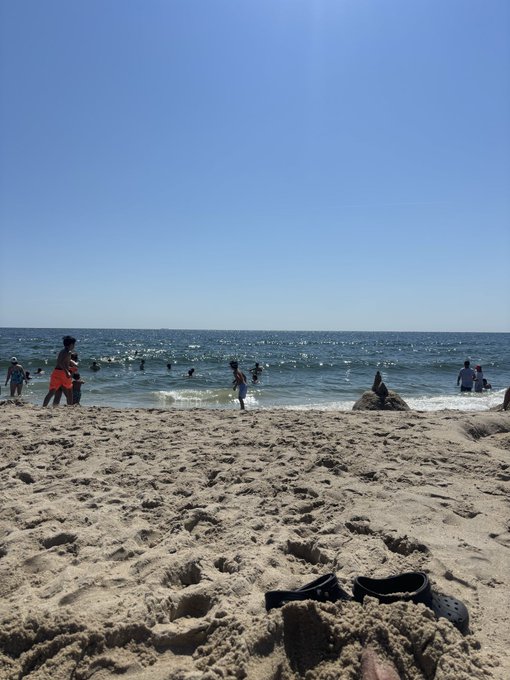 A sunny beach scene with several people wading and swimming in the ocean under a clear blue sky, with a pair of black Crocs half-buried in the sand in the foreground.