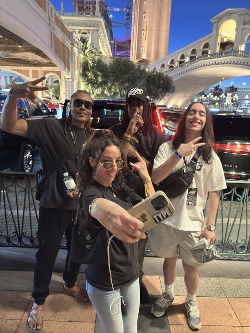 A group selfie of four people posing on a city street at dusk with luxury cars in the background; the person holding the phone has a phone case with "TPXE" visible.
