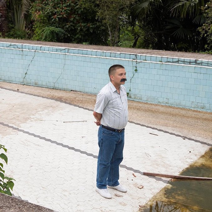Photograph of a man with a mustache wearing a white shirt and blue jeans standing by an empty tiled swimming pool.