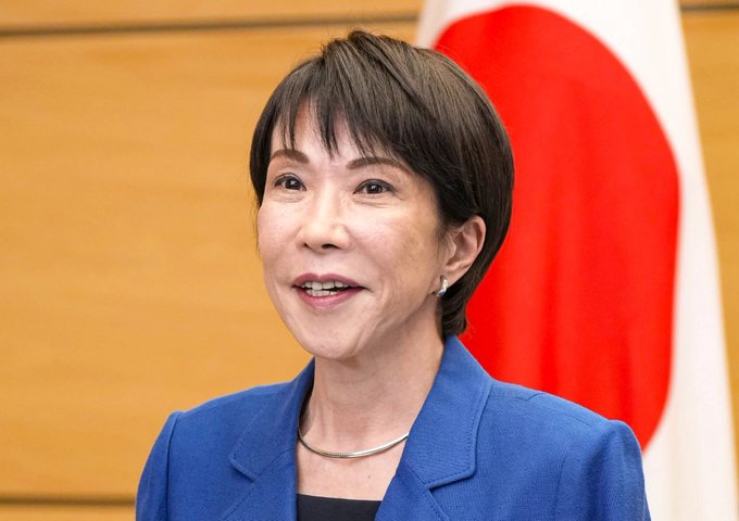 The image shows a woman wearing a blue jacket with her face turned towards the camera. The background features a flag of Japan, which has red and white colors. The caption reads "Why Did Markets Fall? Everyone'S Talking About America. The Real Answer Is in Japan. Yesterday, strong employment data came out of America. Nearly twice what was expected. Everyone pinned the drop on that. Stock market down, gold down, Bitcoin down".