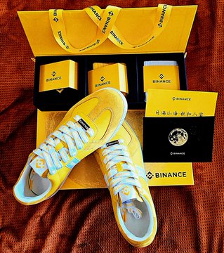 A pair of yellow and white sneakers displayed with matching Binance-branded boxes, socks, and promotional cards on a red fabric background.