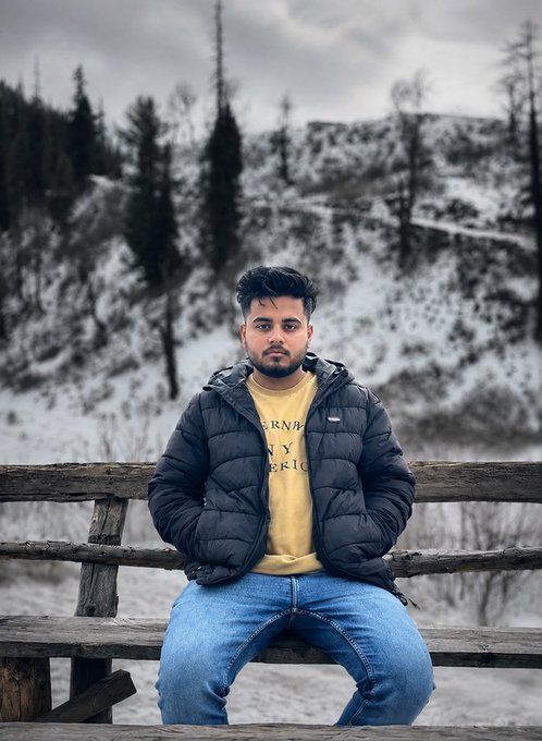 A young man wearing a black puffer jacket over a yellow Burberry t-shirt and blue jeans sits on a wooden bench with a snowy mountain landscape in the background.