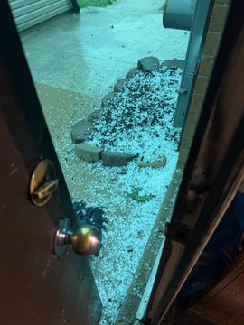Vertical video screenshot showing a shattered glass door or window from the inside, with broken glass pieces scattered on the floor outside.