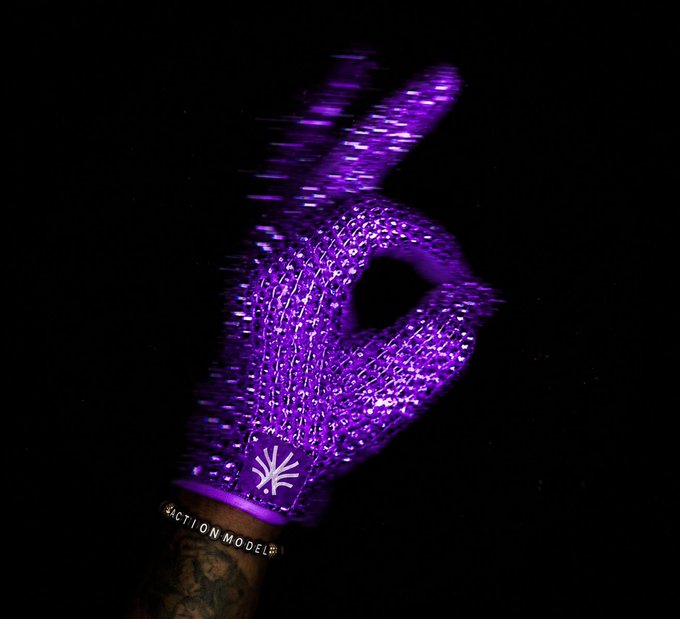 A close-up of a tattooed hand wearing a glowing purple LED light-up glove forming an 'OK' hand gesture against a black background, with 'FRACTION MODEL' text visible on the wrist.