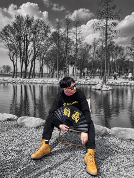 A person wearing a black sweatshirt with gold Chinese characters, a red mask, sunglasses, and tan work boots sits on a suitcase by a pond in a black-and-white photograph with selective color.
