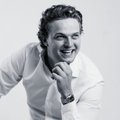 A black-and-white portrait of a smiling man with short hair wearing a white shirt, with his hand resting near his chin, and an Apple Watch visible on his wrist.