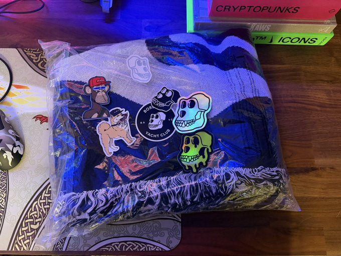 A folded clothing item wrapped in plastic with Bored Ape Yacht Club-themed patches, including skulls and a monkey, sits on a desk next to a CryptoPunks book.