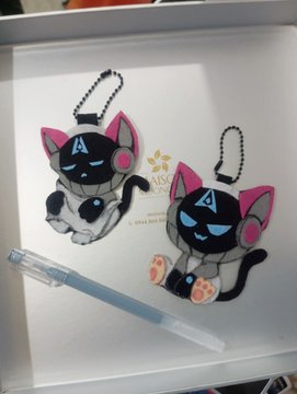 Two 'Valorant' style robot cat keychains with pink ears and a triangular 'A' symbol on their foreheads are displayed in a white box, accompanied by a pen.