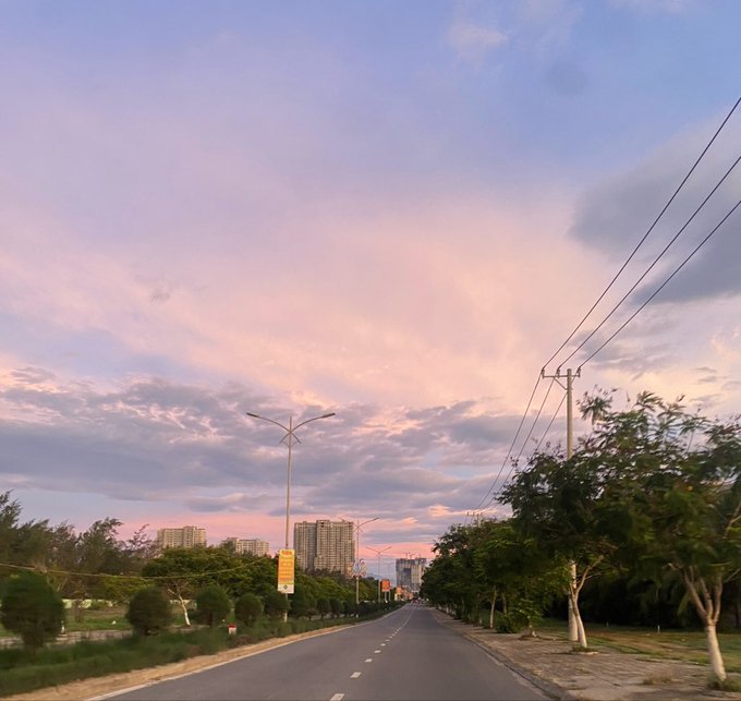 A scenic view of an empty road at sunset or sunrise, with pink and blue clouds, streetlights, power lines, and buildings in the distance.
