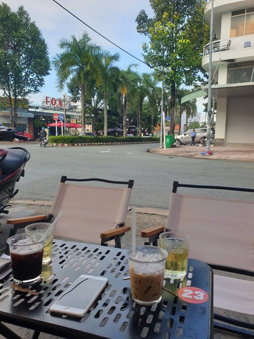 An outdoor cafe scene shows a metal table with iced coffee drinks and a smartphone on it, with chairs, palm trees, and a street view in the background.