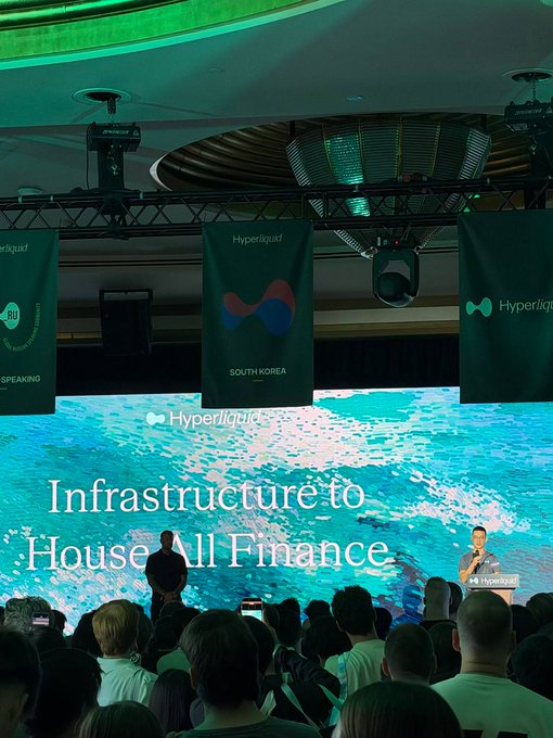 A photograph of a Hyperliquid conference presentation with the slogan 'Infrastructure to House All Finance,' showing a speaker at a podium on stage in front of a large digital screen displaying an ocean scene.