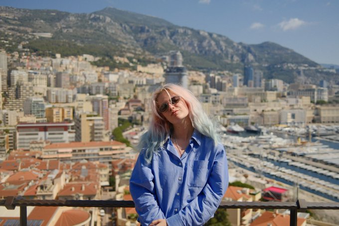 A young woman with pink and blue hair and sunglasses stands at a scenic overlook with a sprawling cityscape, marina filled with yachts, and mountains in the background, likely Monaco.