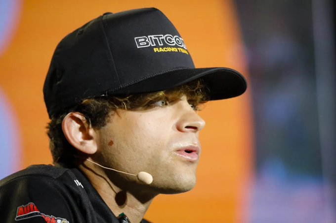 A young man wearing a black 'BITCOIN RACING TEAM' cap and a microphone headset, appearing to speak at an event.
