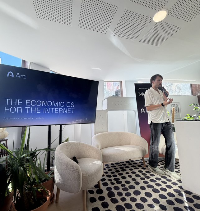 A man speaks into a microphone at an event titled "THE ECONOMICS OF THE INTERNET" hosted by Arc, standing in a modern room with cream-colored chairs.