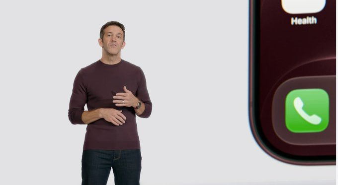 A man in a maroon sweater stands on a stage presenting, with a large image of an iPhone screen showing the Phone and Health app icons visible on the right.