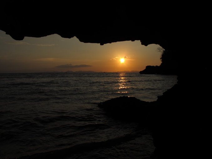 A scenic photograph of a sunset over the ocean, viewed from inside a dark cave or rocky overhang, with the bright sun reflecting on the rippling water and silhouetted cliffs in the distance.