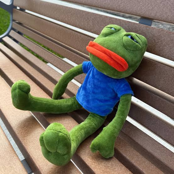 A sad Pepe the Frog plush toy wearing a blue shirt slumps lifelessly on a wooden park bench, limbs splayed.