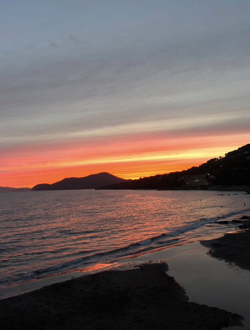 A sunset over the ocean paints the sky orange and pink, with mountains silhouetted along the coastline.