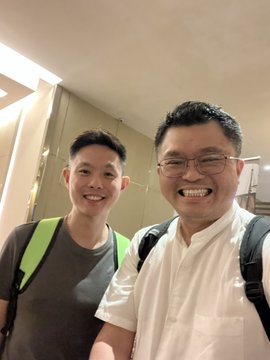 A selfie of two smiling men, one wearing glasses and a white shirt with a backpack strap, and the other wearing a grey t-shirt with a bright green backpack strap.
