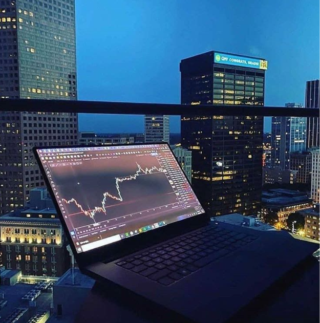 A laptop displays stock market charts on a balcony overlooking a city skyline at night, featuring a building with the text "OPT CONGRATS, GRAD!". This image visually represents the post's theme of market activity continuing even when the city sleeps.