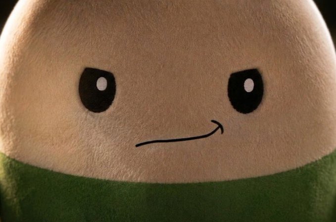 Close-up of a plush character with a round white face, black eyes, and a simple curved smile drawn on, wearing a green collar or scarf.