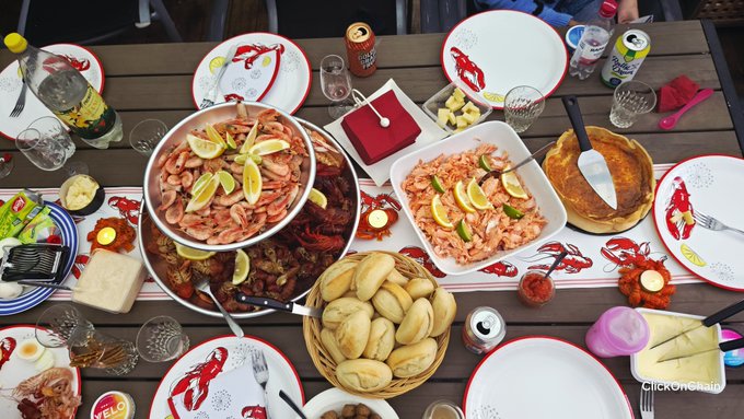 A festive spread of Swedish seafood—including bowls of shrimp garnished with lemon, pickled herring, and a basket of bread—set among lobster-printed plates and drinks on a wooden cabin table, marking the final weekend day before returning to trading routines.