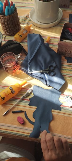 Craft workspace with a Sonic the Hedgehog silhouette cut from blue fabric, surrounded by glue bottles, scissors, a pen, eraser, a glass of water, and a basket of markers.