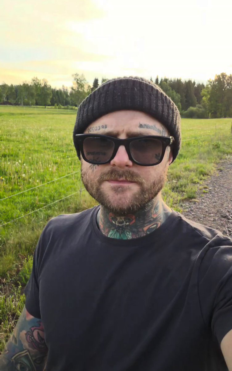 The image features a man wearing sunglasses, a black hat, and a t-shirt with tattoos. He has a beard and appears to be posing for a photo. The background of the image shows a field or pasture with trees, giving off a peaceful atmosphere. Below the main subject, there is a post that reads "Good night and Gn X Join us in the celebration for the 1 year anniversary of @SolOnChain tommorow Set your reminders in the post below". This suggests that the man might be celebrating an event related to the Sol On Chain platform or a similar project.