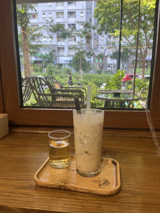 An iced latte and water sit on a wooden Starbucks tray by a window overlooking a city park, offering a quiet moment of respite. The image evokes the contemplative, somewhat melancholic mood of the post—finding small comforts like a coffee break while weathering the slow, drawn-out downtrend in crypto markets.