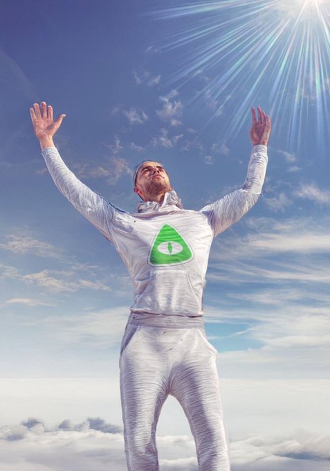 A man in a silver outfit with a green triangular symbol stands with arms raised toward the sun, surrounded by clouds and blue sky.