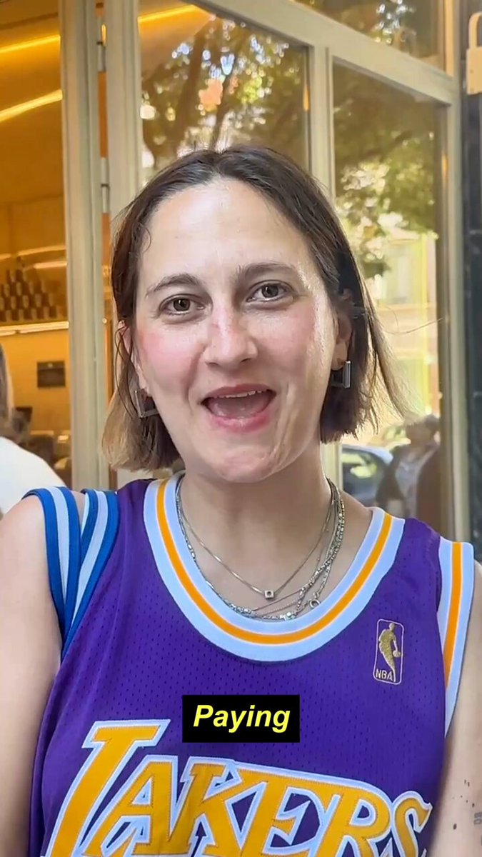 The image features a woman wearing a purple and yellow basketball jersey that says "Lakers" on it. She has short brown hair and is smiling at the camera. The background of the photo shows a window, which might be part of a store or restaurant. There are also other people visible in the background, but they are not the main focus of the image.