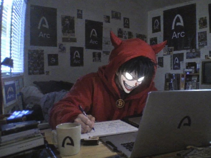 A stylized edited image of a person wearing a red devil hoodie with glowing white eyes and a sinister grin, working at a desk covered in 'Arc' branding on stickers, a laptop, and a coffee mug.