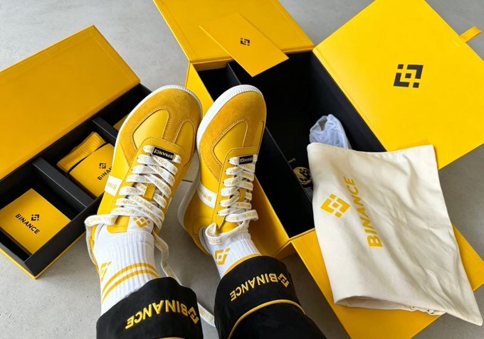 A pair of yellow Binance-branded sneakers with white laces and socks are displayed on top of their yellow shoebox packaging, accompanied by a Binance dust bag.