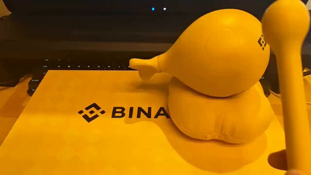The image features a yellow rubber duck sitting on top of a computer keyboard, with the word "binance" written on the keyboard. The rubber duck appears to be made out of foam or plastic material, giving it a playful appearance. The background is blurred, drawing attention to the rubber duck and its position on the keyboard.