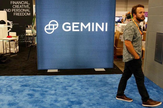 A man walks past a large blue 'GEMINI' display banner at a convention, with a sign partially visible reading 'FINANCIAL, CREATIVE, AND PERSONAL FREEDOM'.