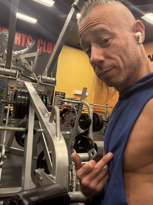 An older, lean man with long gray hair and AirPods stands in a gym, flexing slightly while looking at the camera.